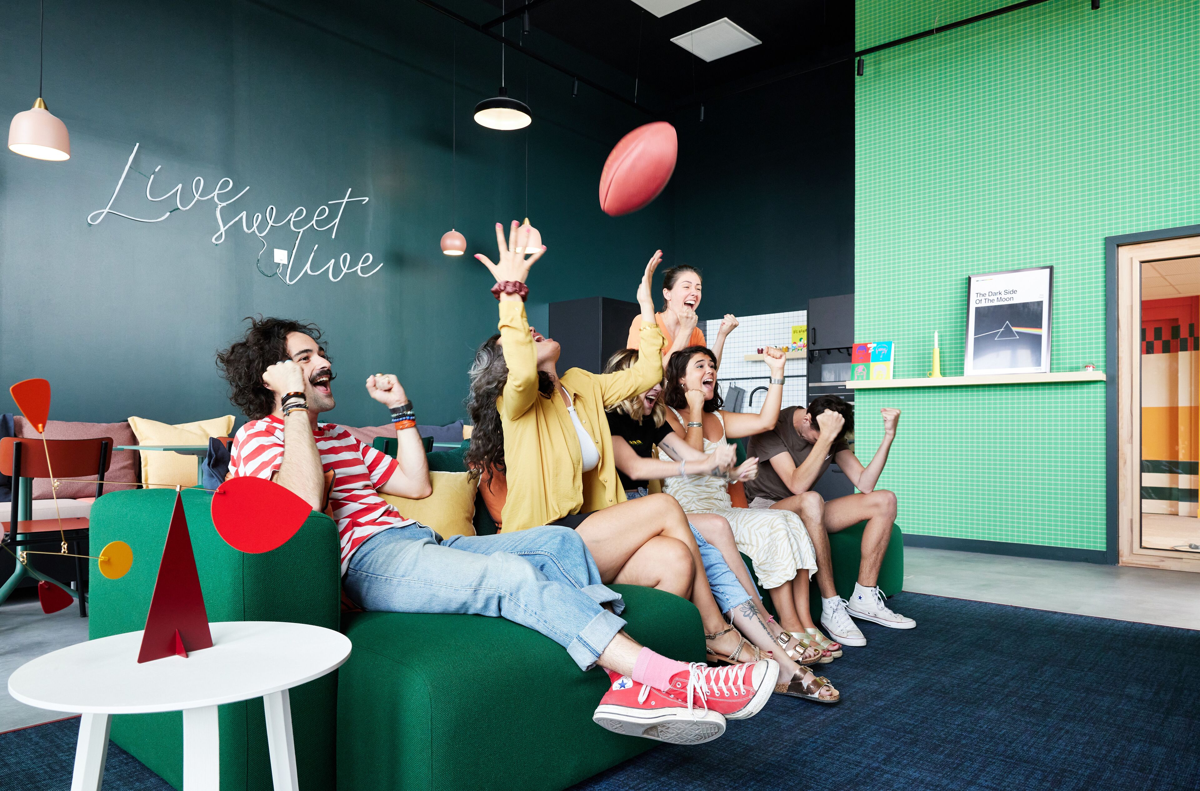Een groep mensen zit op een groene bank in een kamer met een groene wand en een bord waarop staat: Live sweet live.