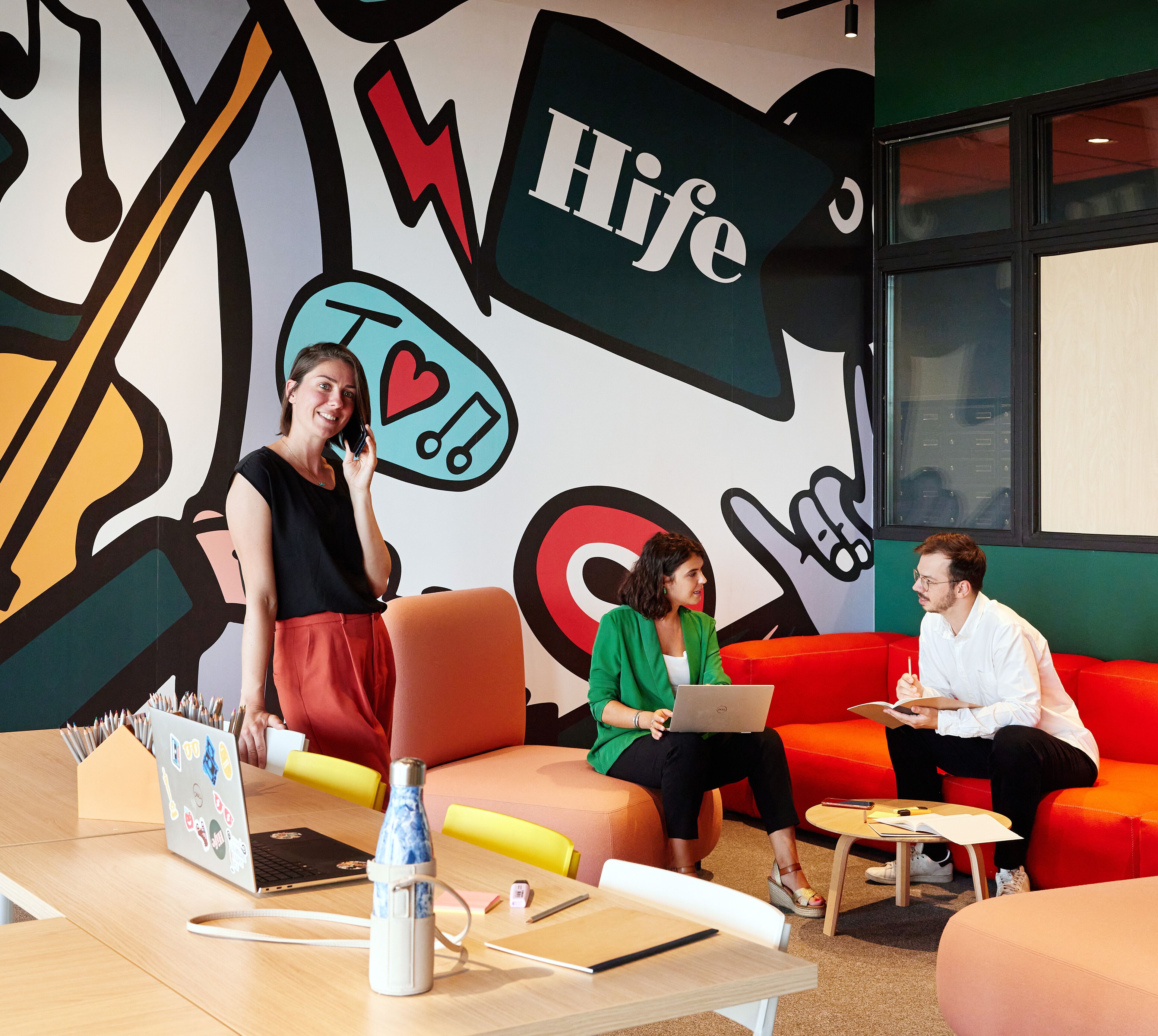 A modern office space with colorful artwork on the walls, a laptop on a table, and people engaged in conversation.