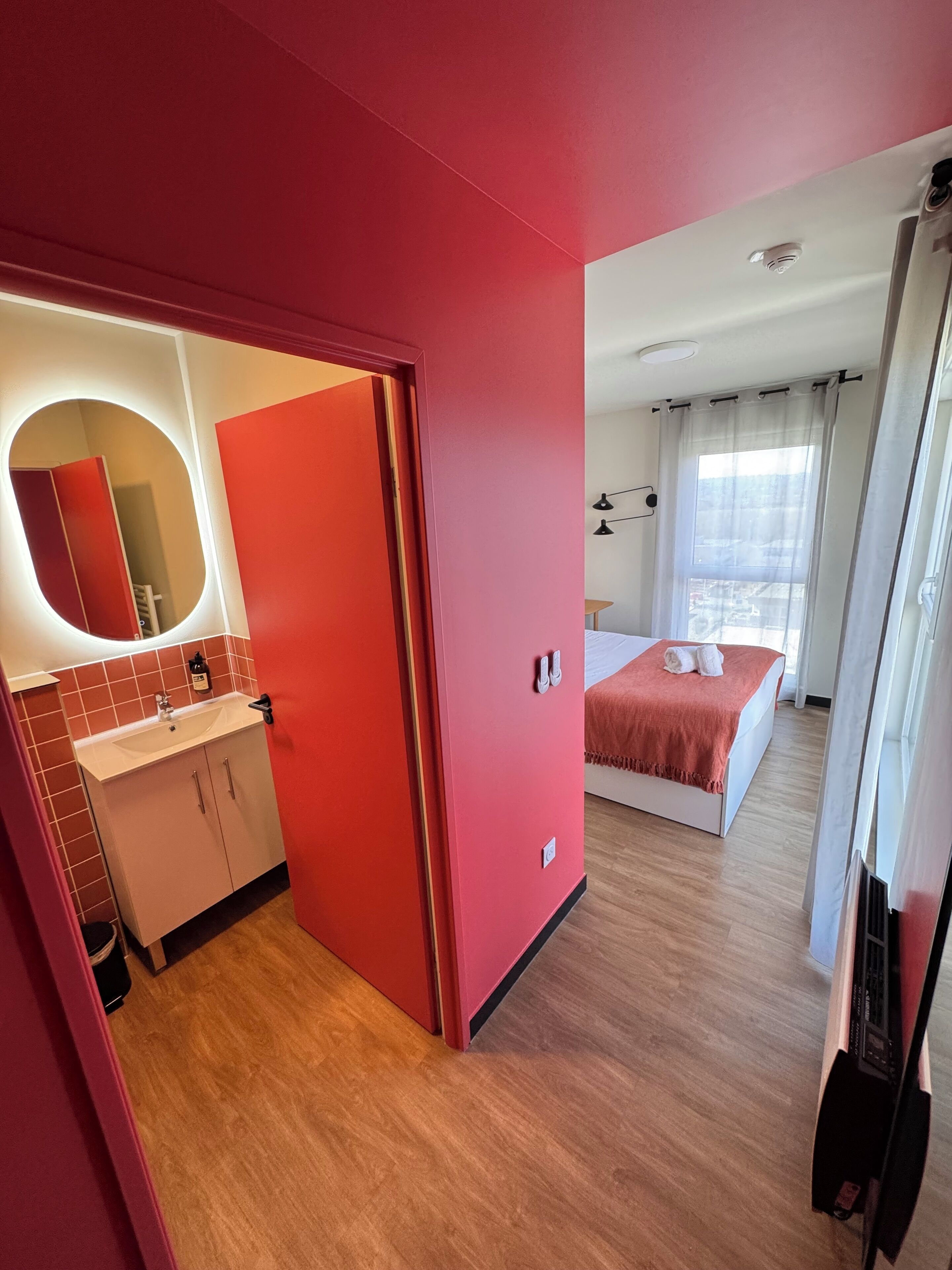 A modern hotel room with a red accent wall, a large bed, a bathroom with a mirror and sink, and a view of the city through the window.