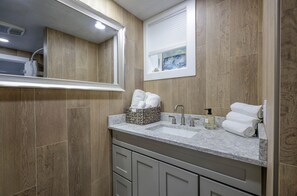 A bathroom with a wooden wall, a sink with a marble countertop, a mirror, and a window with blinds. - Lower Level Apartment Close to Town and Beaches (2314) (Brewster)