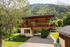 Un chalet de madera de dos pisos con balcón, rodeado de un jardín cuidado y con montañas al fondo.