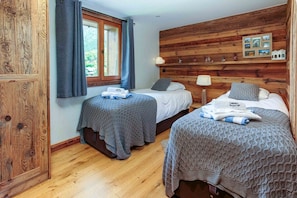A room with two beds, wooden paneling on the walls, a window with curtains, and a small shelf with pictures. - Chalet du Dard, amazing views, Chamonix All Year (Chamonix)