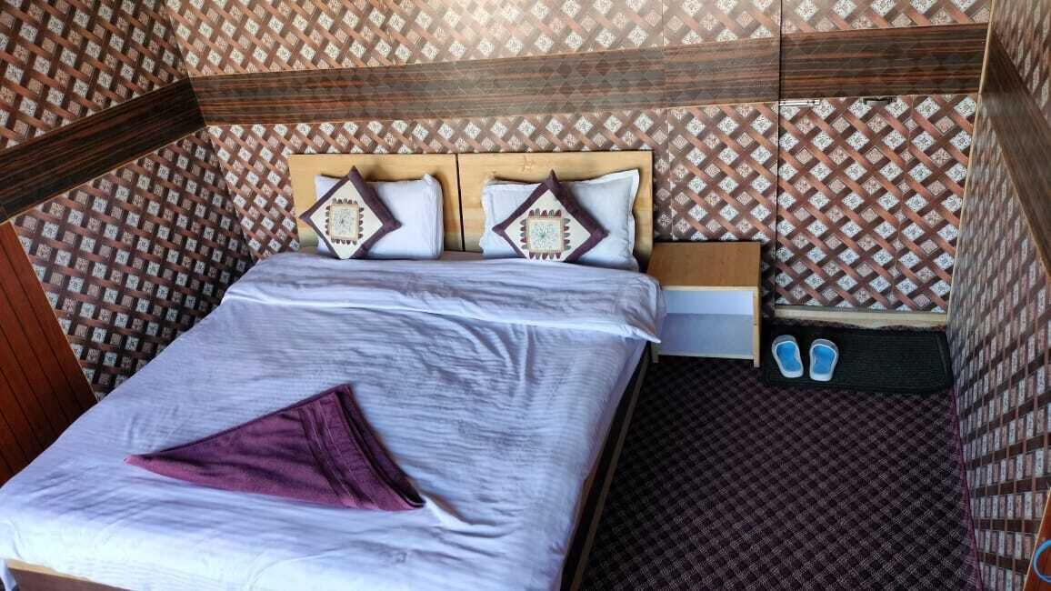 Una cama individual con cabecera de madera, un cojín morado y una mesita de noche con un reloj y una caja.