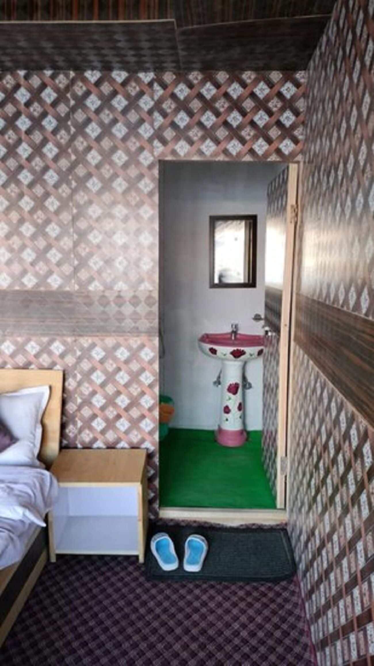 Una habitación pequeña y estrecha con papel tapiz estampado, una cama individual, una mesita de noche, un lavamanos con espejo y un par de zapatos en el suelo.