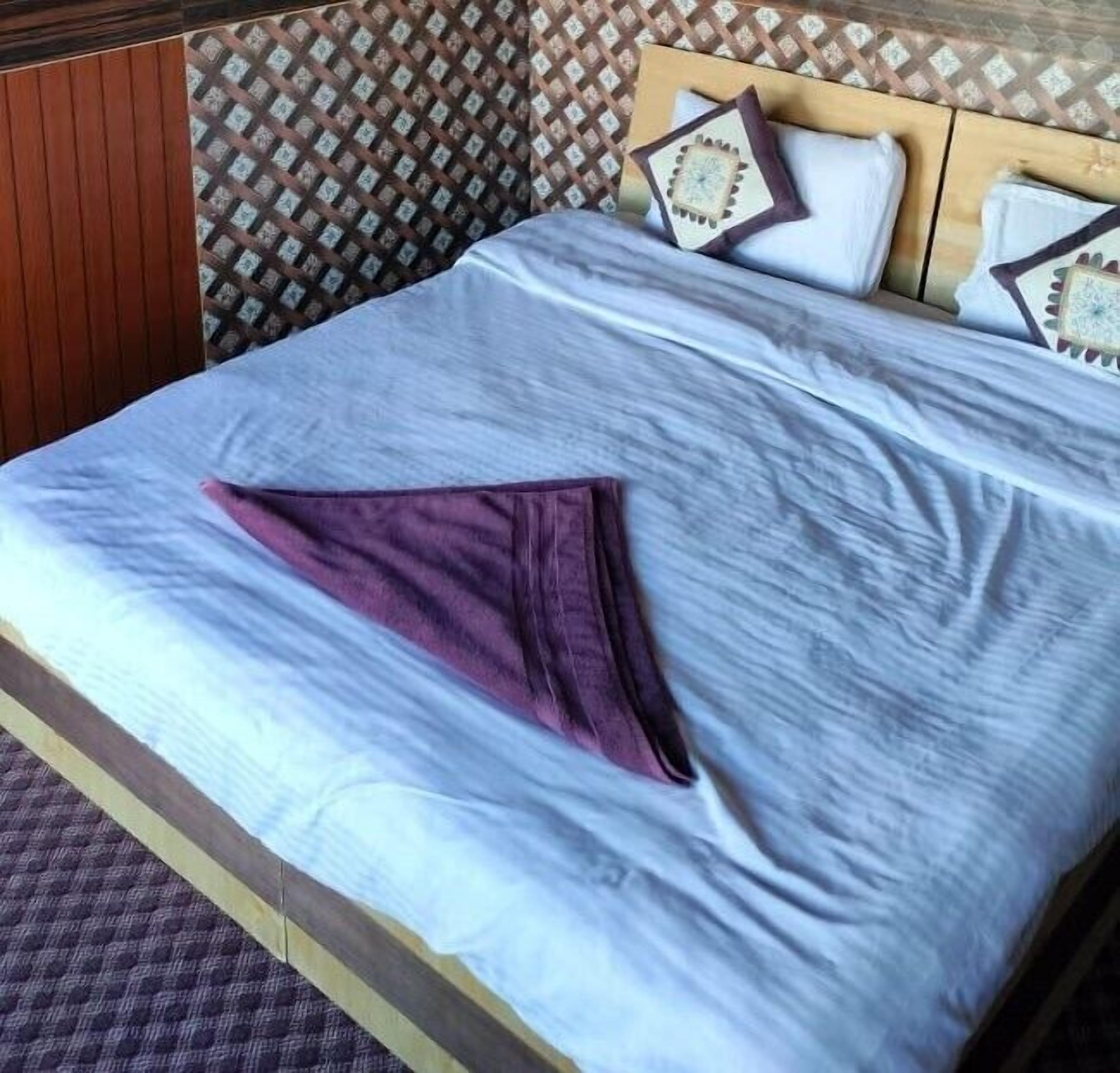 A bed with a blue bedspread, a purple pillow, and a wooden headboard with patterned wallpaper.