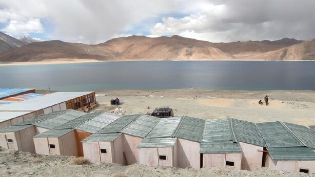 Une rangée de bâtiments simples, à un seul étage, coiffés de toits en métal, situés dans un paysage aride et dégagé, avec une vaste étendue d’eau et des montagnes en arrière-plan.