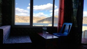 Une pièce dotée d’une grande fenêtre donnant sur un paysage montagneux et un plan d’eau. On y trouve une chaise bleue et une petite table sur laquelle sont posés une bouteille d’eau et des verres.