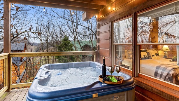 A hot tub with a bottle of wine and glasses, a wooden deck, and a view of trees and buildings. - 3 Primary Suites, Amazing location, Hot Tub (Pigeon Forge)