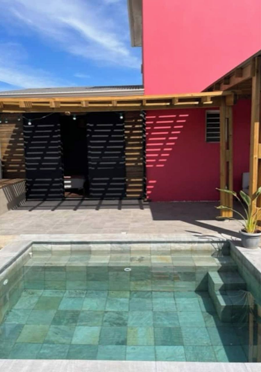A swimming pool with a tiled deck, a red building with a sloped roof, and a wooden pergola structure.