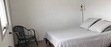 A small, sparsely furnished room with a single bed, a chair, a table, and a hanging light fixture.