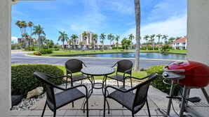 A covered patio with two chairs and a table, a red grill, and a view of a lake with palm trees and buildings in the background.