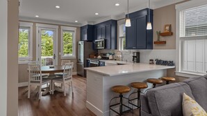 A modern kitchen with dark blue cabinets, a white island with bar stools, and a dining area with white chairs. - Seabrook SeaDragon | Pet Friendly, Hot Tub, 3BR | Sleeps 9, Sunrise Pool (Pacific Beach)
