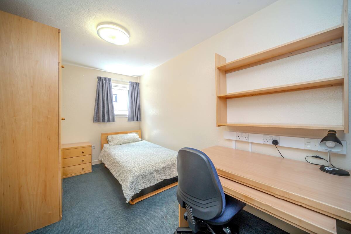 A bedroom with a bed, a desk, a chair, a wardrobe, and a shelf.
