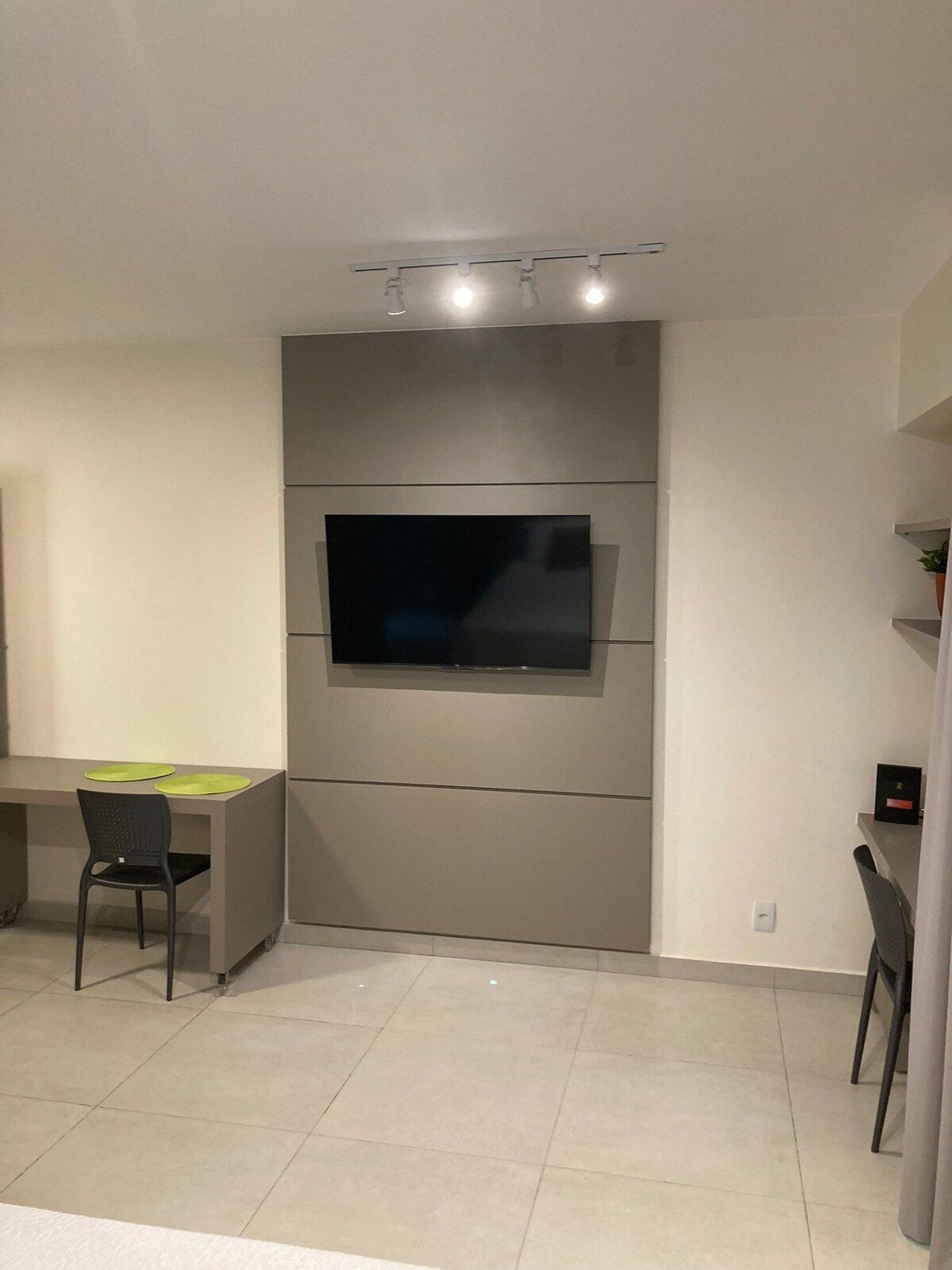 A modern living room with a flat-screen TV mounted on a wall, a desk with a chair, and a small plant on a shelf.