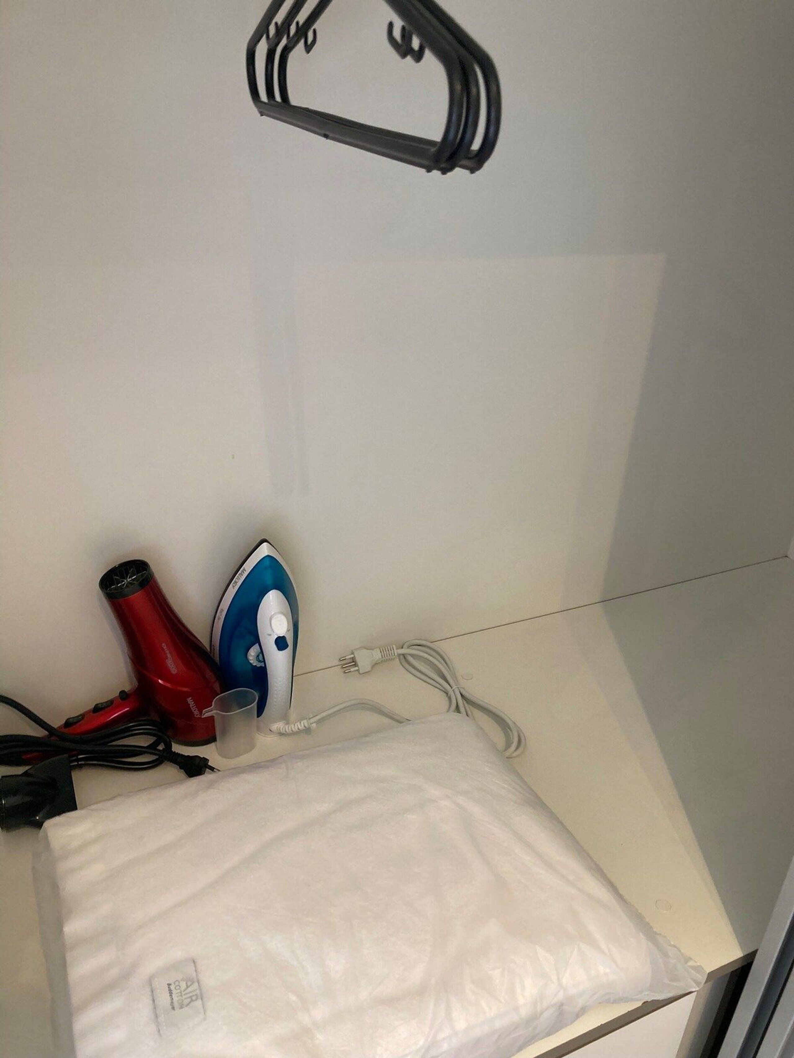 Ein Zimmer mit weißer Wand, einem hängenden Kleiderständer, einem roten Föhn, einem blau-weißen Bügeleisen und einer weißen Plastiktüte auf einer weißen Fläche.
