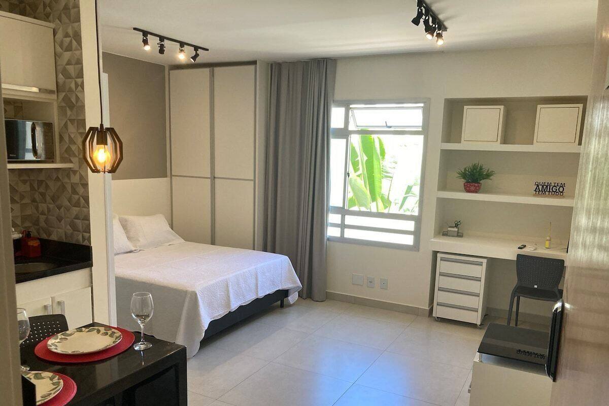 A modern hotel room with a bed, a desk, a chair, a small table with a wine glass, and a window with curtains.