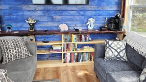 Un salon chaleureux avec un mur en bois bleu, un téléviseur à écran plat, une bibliothèque remplie de livres, deux canapés gris et une machine à café.