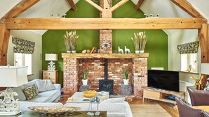 A cozy living room with a brick fireplace, wooden beams, a television, and a sofa. - Hollington Barns - Luxury Cottages (Ashbourne)