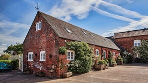 A red brick building with a sloped roof, multiple windows, and a small garden area with potted plants and flowers. - Hollington Barns - Luxury Cottages (Ashbourne)