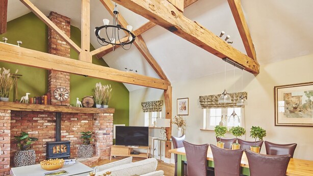 A cozy living room with exposed wooden beams, a brick fireplace, and a dining area with modern furniture. - Hollington Barns - Luxury Cottages (Ashbourne)