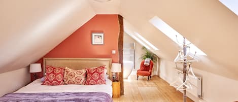 Un dormitorio con techo inclinado, una cama con almohadillas estampadas, una silla roja, una lámpara de pie y una araña.
