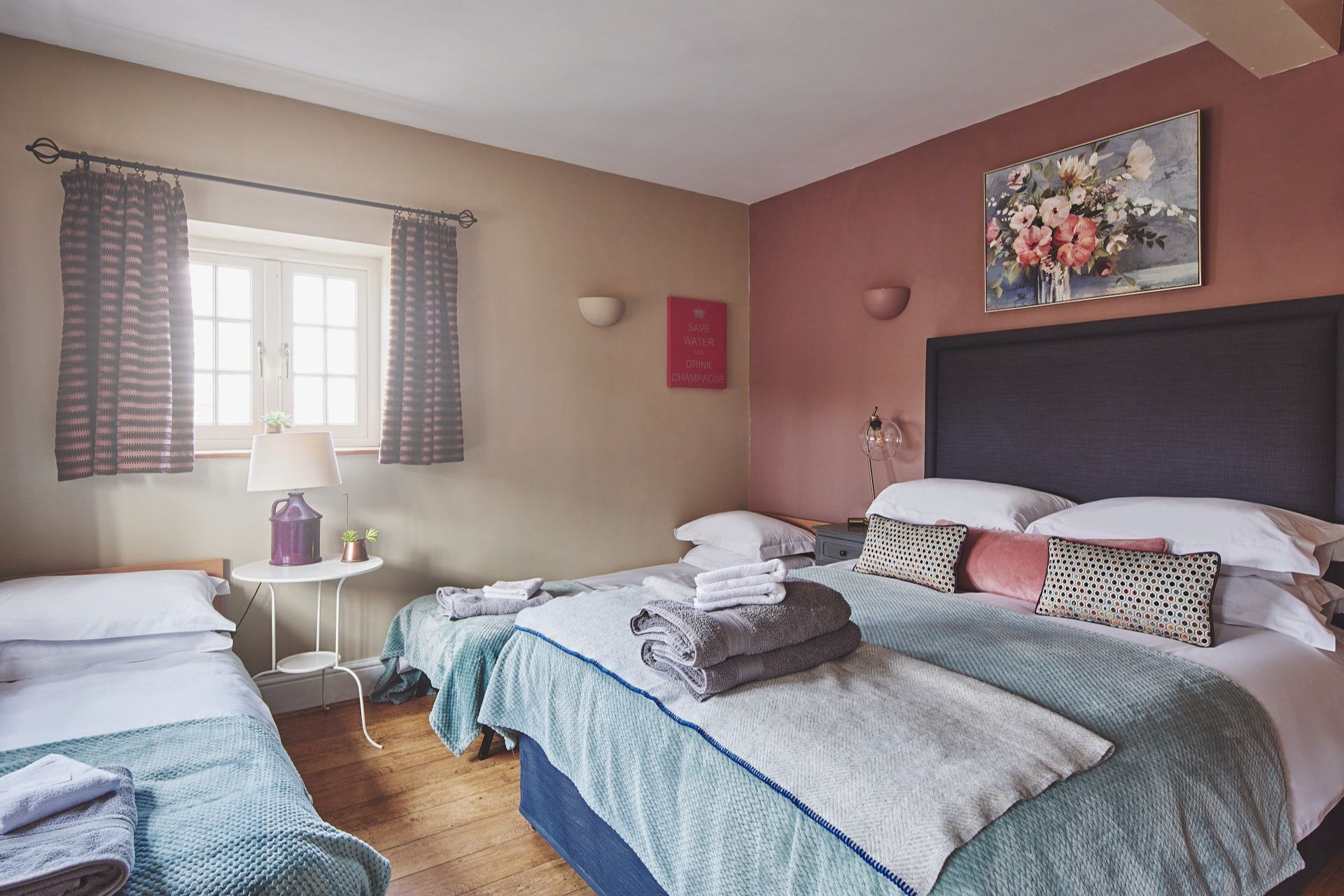A bedroom with two beds, a small table, a lamp, a potted plant, and a painting on the wall.