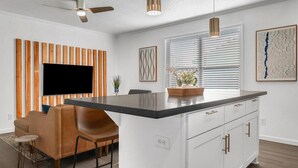 A modern kitchen with a central island, white cabinetry, and a flat-screen TV mounted on a wooden panel wall. - BethyKay: 2 Bedroom, Walk to A&M, Comfy King Beds (Bryan)