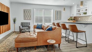 A modern living room with a leather sofa, a grey armchair, and a coffee table. There is a kitchen area with a bar and stools. - BethyKay: 2 Bedroom, Walk to A&M, Comfy King Beds (Bryan)