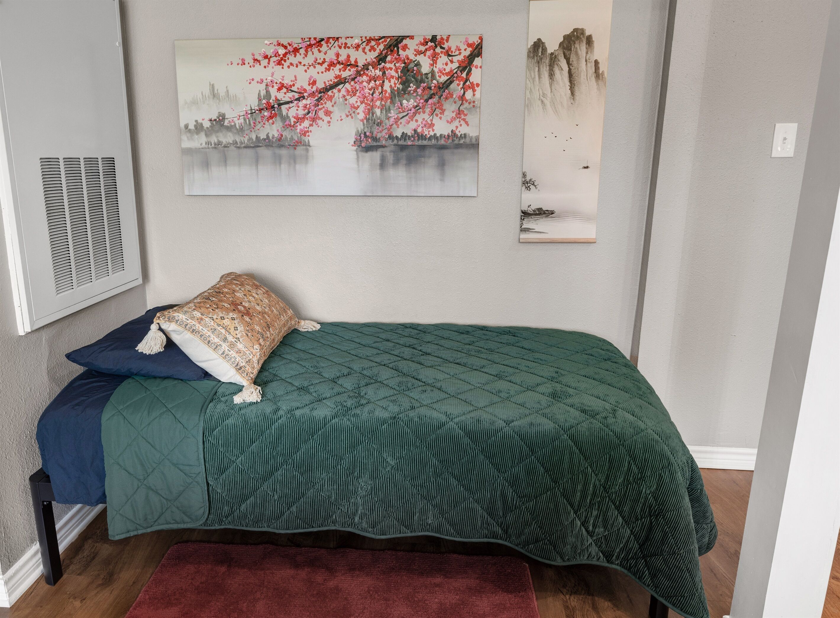 A bedroom with a bed, pillows, a quilted green bedspread, a red rug, and two wall-mounted paintings.