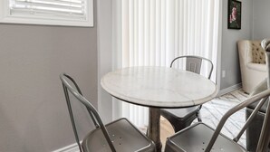 A dining area with a round table and four chairs, a window with blinds, and a wall-mounted picture. - Blossom: Ground Floor, Comfy King Bed (Bryan)