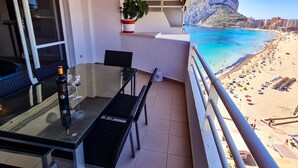 A balcony with a glass table, chairs, a bottle, and a glass of wine. A potted plant is on the wall. The balcony overlooks a beach with people and umbrellas.