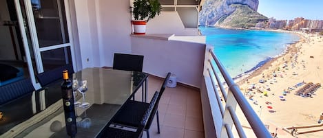 Un balcón con una mesa de vidrio, sillas, una botella y una copa de vino. Hay una planta en maceta en la pared. El balcón da a una playa con gente y sombrillas.
