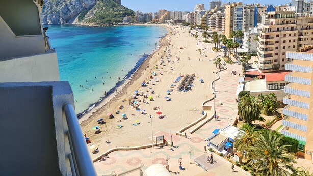 A beach with a rocky mountain in the background, a promenade with umbrellas, and a cityscape with buildings.