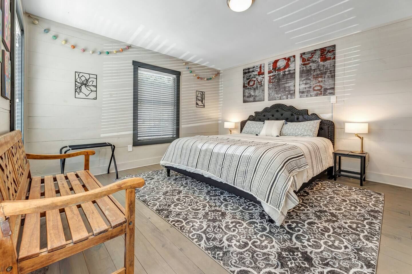 Une chambre à coucher comprenant un lit, deux tables de chevet, un banc, une fenêtre avec des stores et des œuvres d’art murales.