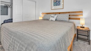 Un dormitorio con una cama grande, cabecera de madera, dos mesitas de noche con lámparas, un armario y un cuadro enmarcado en la pared.
