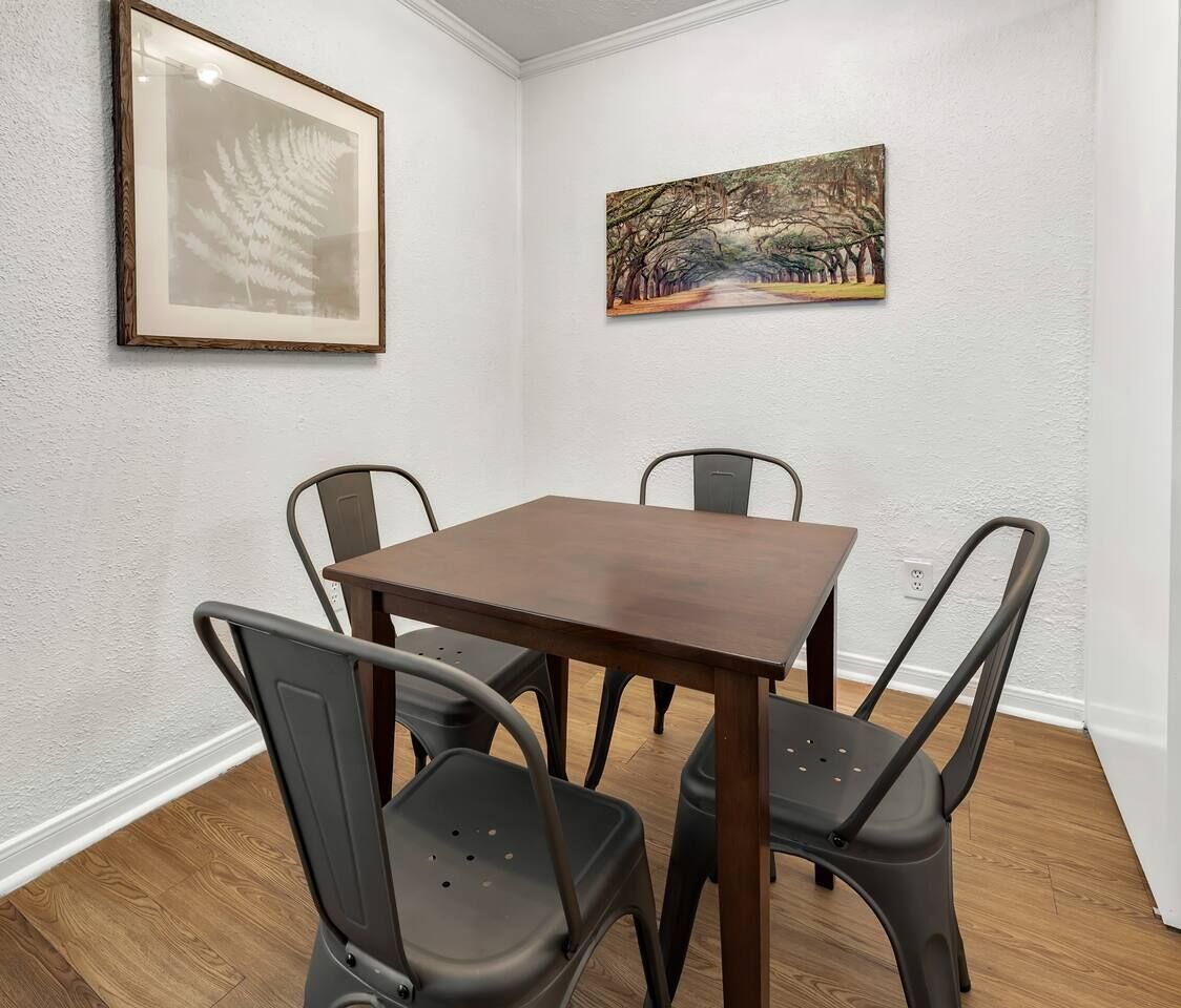 Un coin repas avec une table en bois et quatre chaises, des œuvres d’art encadrées accrochées aux murs et un mur blanc à la texture particulière.