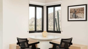 A room with a round table, two black chairs, a vase, a framed picture, and a window with a view of cacti. - Sacred Haven By Homestead Modern (Joshua Tree)