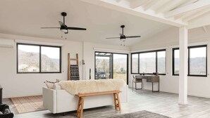 A spacious room with a white sofa, a wooden bench, a desk, and two ceiling fans. Large windows offer views of the outdoors. - Sacred Haven By Homestead Modern (Joshua Tree)