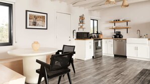 A modern kitchen with white cabinetry, a dining area with a round table and chairs, and a large window providing natural light. - Sacred Haven By Homestead Modern (Joshua Tree)