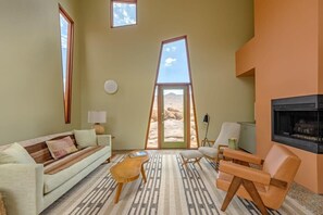 A living room with a sofa, chairs, a coffee table, a fireplace, and a large window with a view of the desert. - Monument House by Homestead Modern (Joshua Tree)