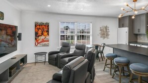 A modern living room with a flat-screen TV, grey leather sofas, a dining area with a table and chairs, and a kitchen with grey cabinets and a bar stool area. - Cinema: 2 Bedroom, Walk to A&M, Comfy King Beds (Bryan)