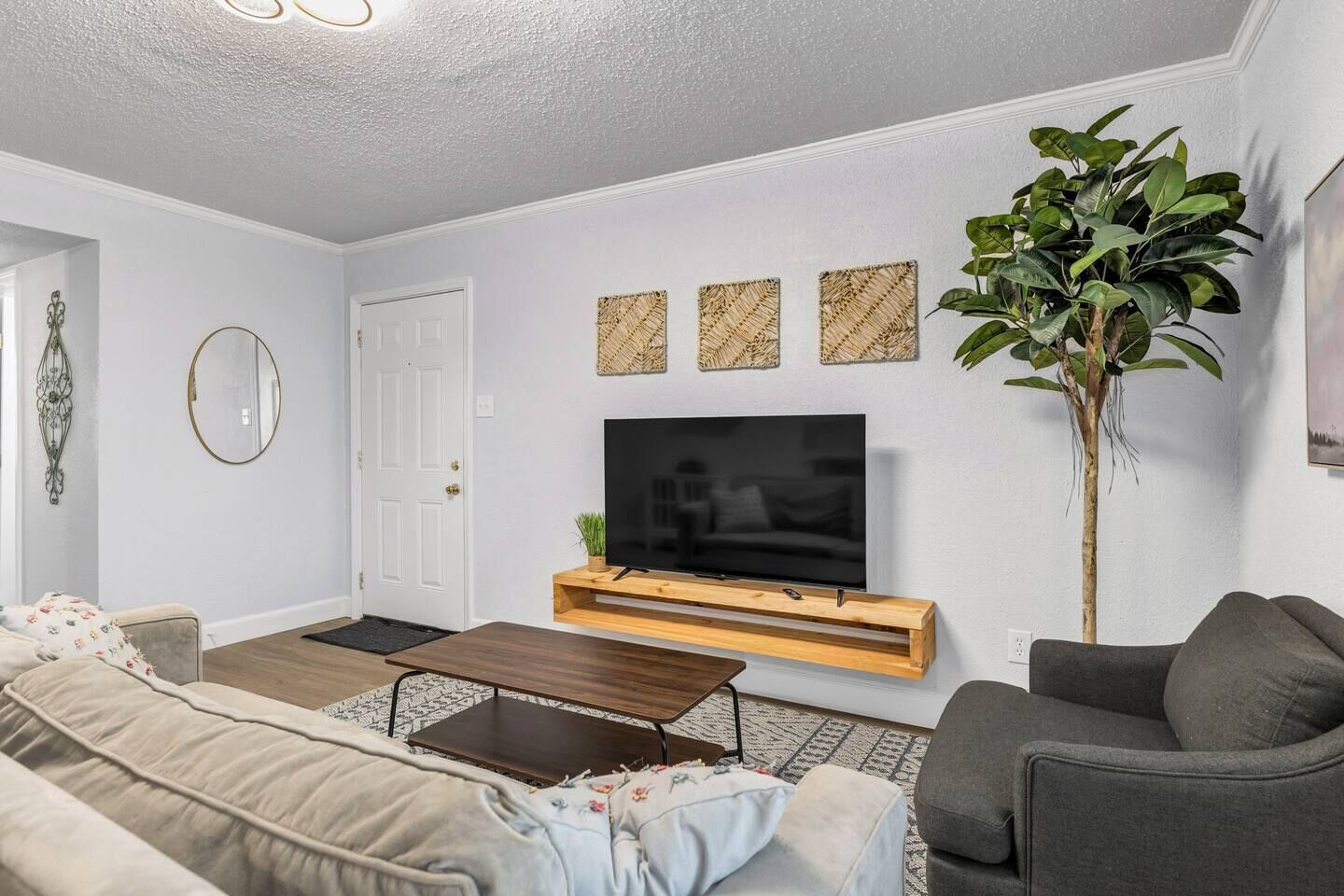 A living room with a flat-screen TV on a wooden stand, a grey sofa, a dark coffee table, and a large plant.