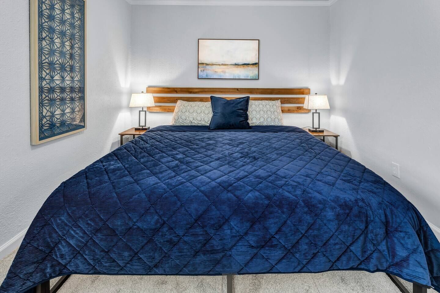 A bedroom with a large bed, two bedside tables with lamps, a wooden headboard, a wall-mounted artwork, and a decorative panel on the left wall.