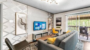 A modern living room with a grey sofa, a flat-screen TV, and a unique wall design. - Denali: 2 Bedroom, Walk to A&M, Comfy King Beds (Bryan)
