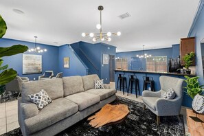 A modern living room with a grey sofa, a wooden coffee table, and a black and white rug. There is a kitchen area with a blue countertop and bar stools. - Comfy King Beds, Close to A&M (College Station)