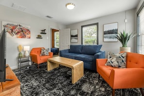 A modern living room with a blue sofa, orange armchairs, and a wooden coffee table. There are framed pictures on the walls and a potted plant by the window.