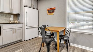 Een kleine keuken met witte kasten, een witte koelkast, een houten eettafel met vier zwarte stoelen, en een raam met jaloezieën.