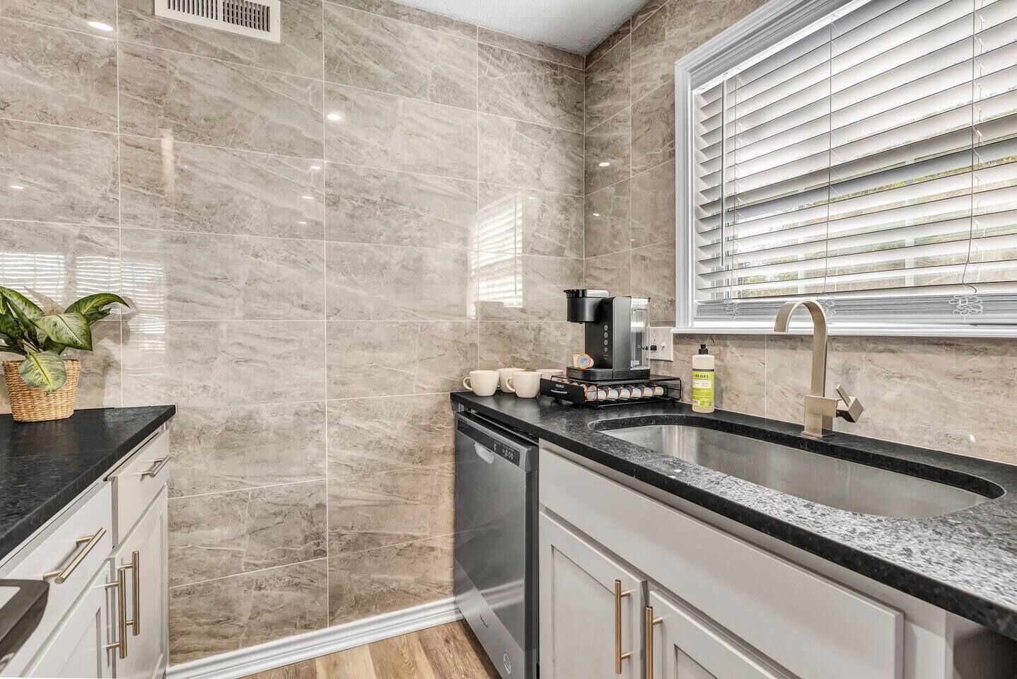 A modern kitchen with marble tiles, a built-in dishwasher, a coffee machine, and a window with blinds.