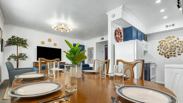 A dining area with a wooden table set for four, surrounded by chairs. There is a potted plant on the table, and a television is visible in the background. - Elise: 2 Bdrm,  Walk to A&M, Comfy King Beds (Bryan)