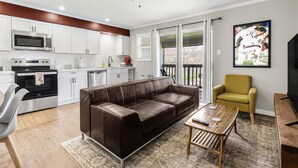 A modern living room with a brown leather sofa, a wooden coffee table, and a yellow armchair. A kitchen with white cabinets and a microwave is visible in the background. - The Zone: Blocks from Texas A&M, Comfy King Bed (Bryan)